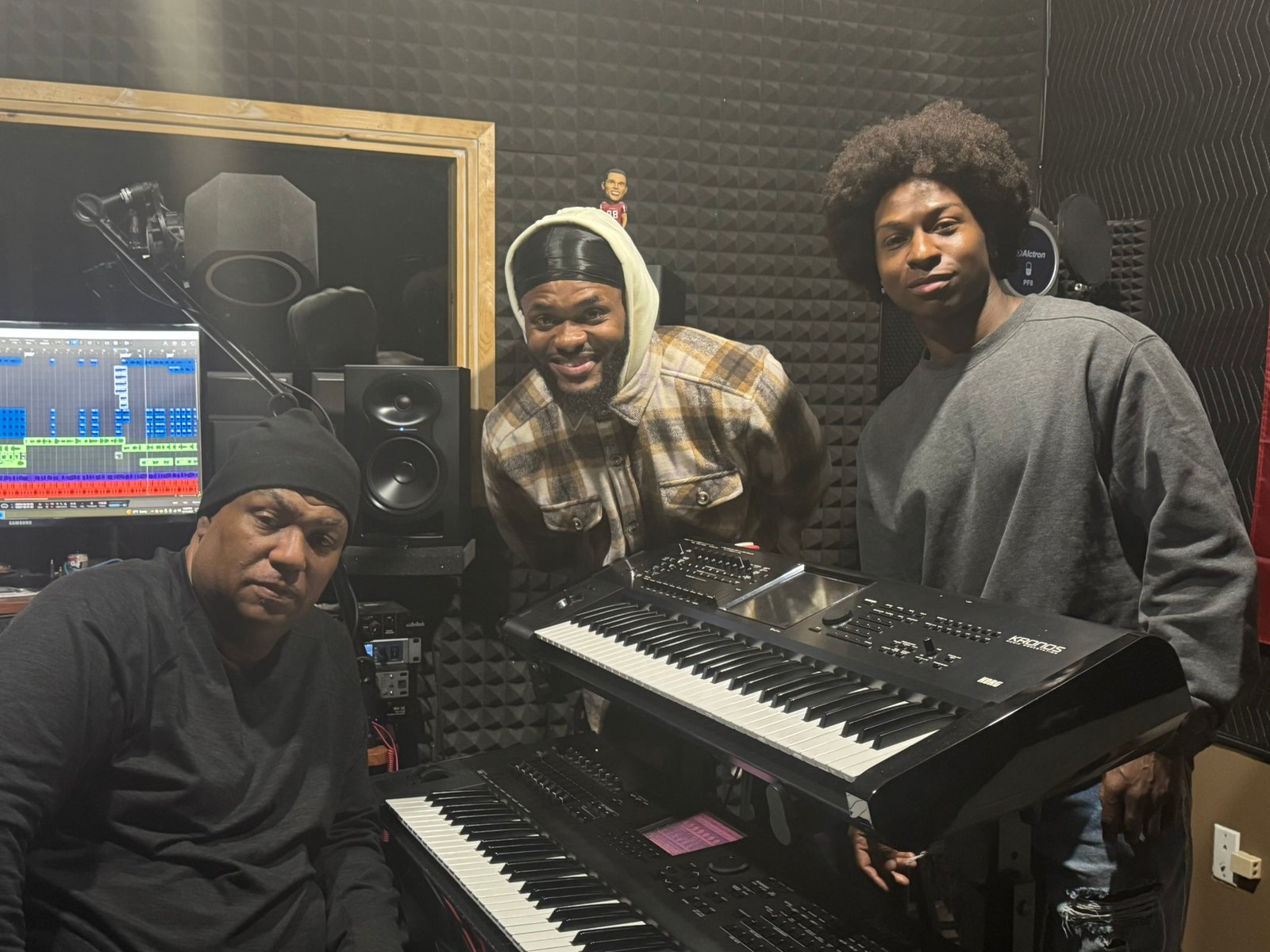Three people together in a recording studio beside keyboards, a microphone, and audio equipment